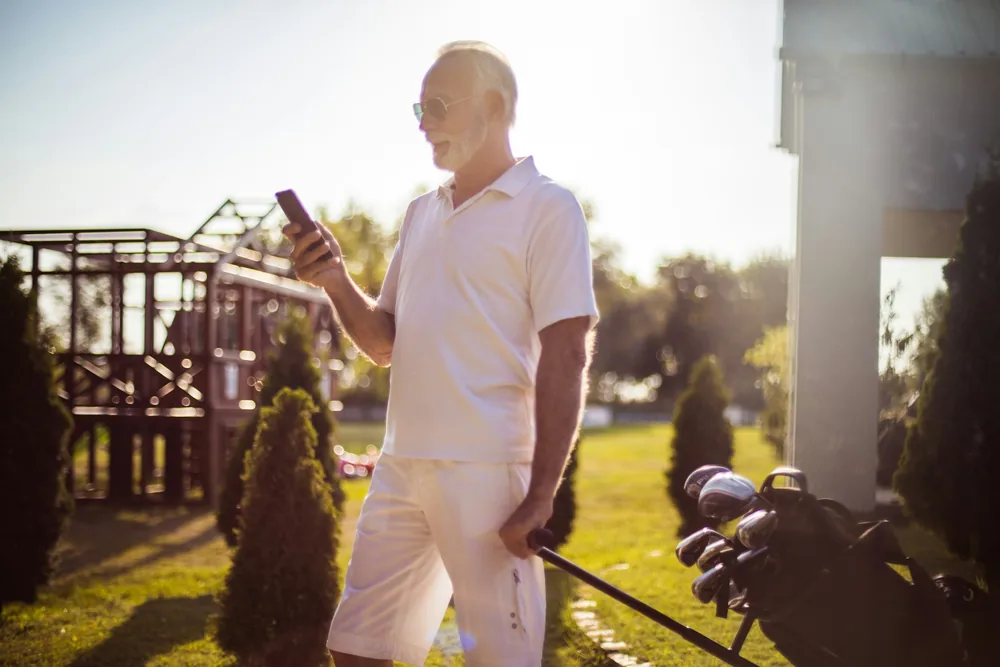 Golfeur senior consultant son smartphone devant le club-house pour retrouver son numéro de licence de golf avant une partie.