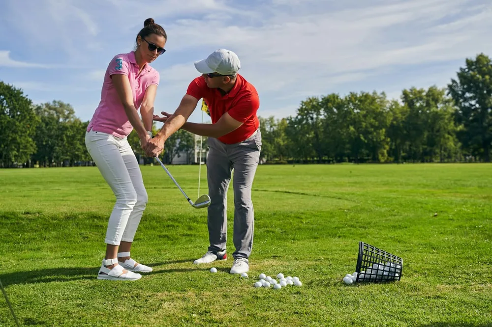 Un enseignant corrige la position des mains d'une golfeuse sur l'herbe, près d'un panier de balles.