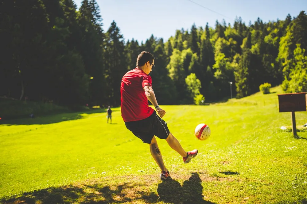 Un joueur en maillot rouge frappe un ballon sur un terrain enherbé bordé d'arbres.